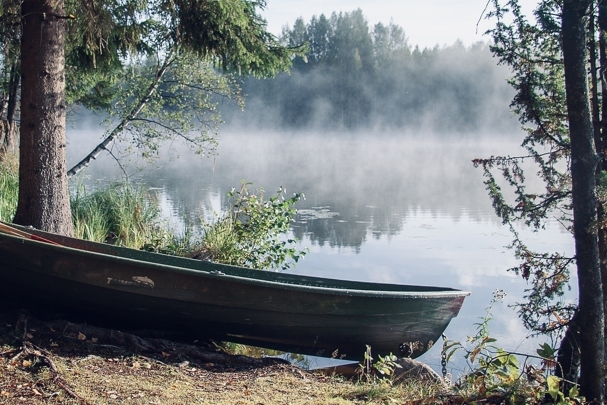 A wooden boat rests on the shore of a misty, forest-lined lake.