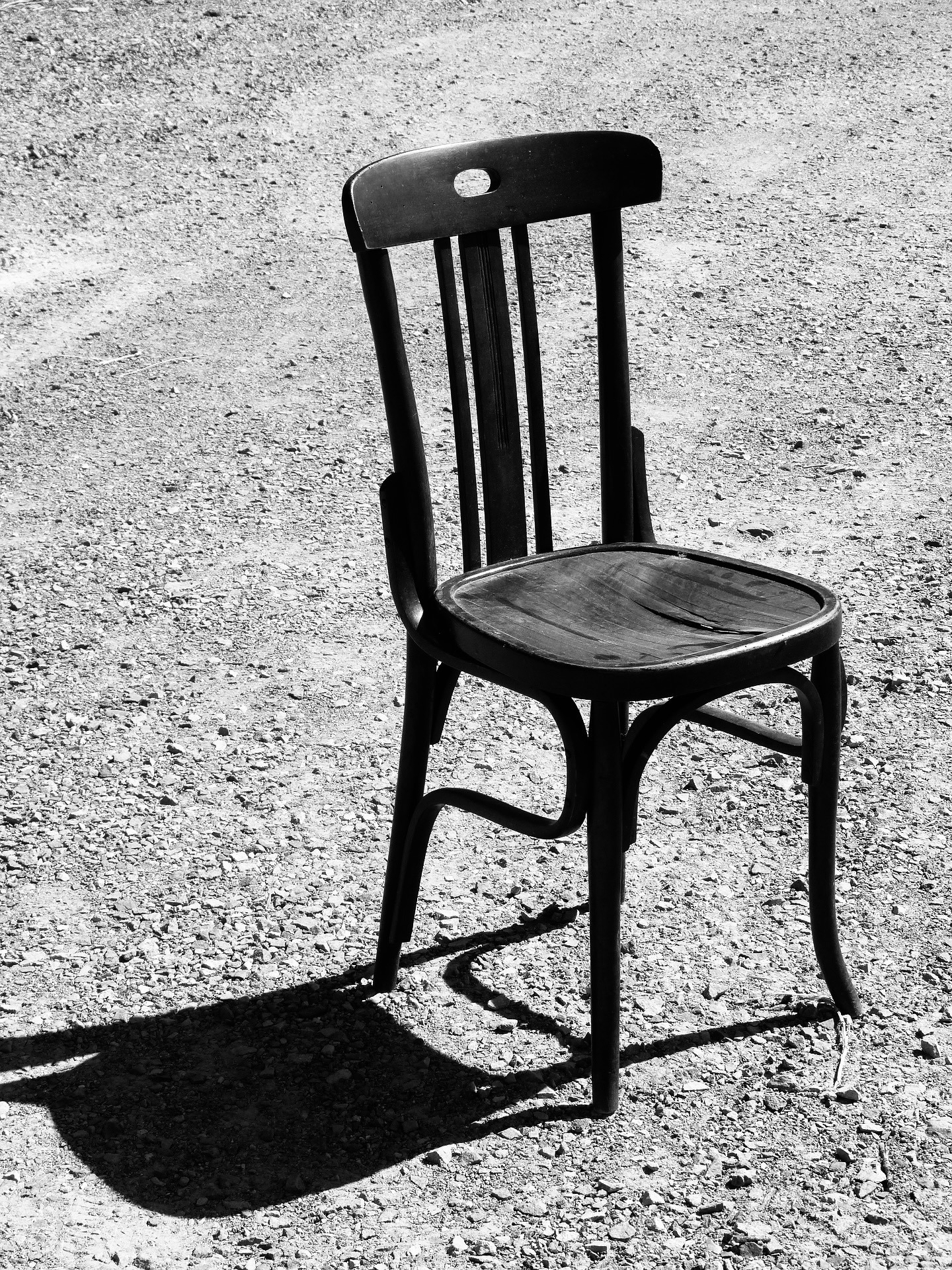 Auto-generated description: A solitary wooden chair casts a shadow on a gravel surface in a black and white photograph.