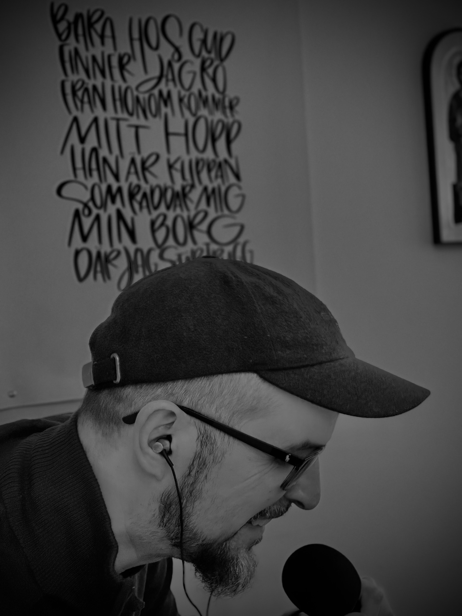 En man med glasögon och keps talar i en mikrofon, med en väggtext i bakgrunden som säger Bara hos Gud finner jag ro, från honom kommer mitt hopp, han är klippan som räddar mig, min borg där jag är trygg.