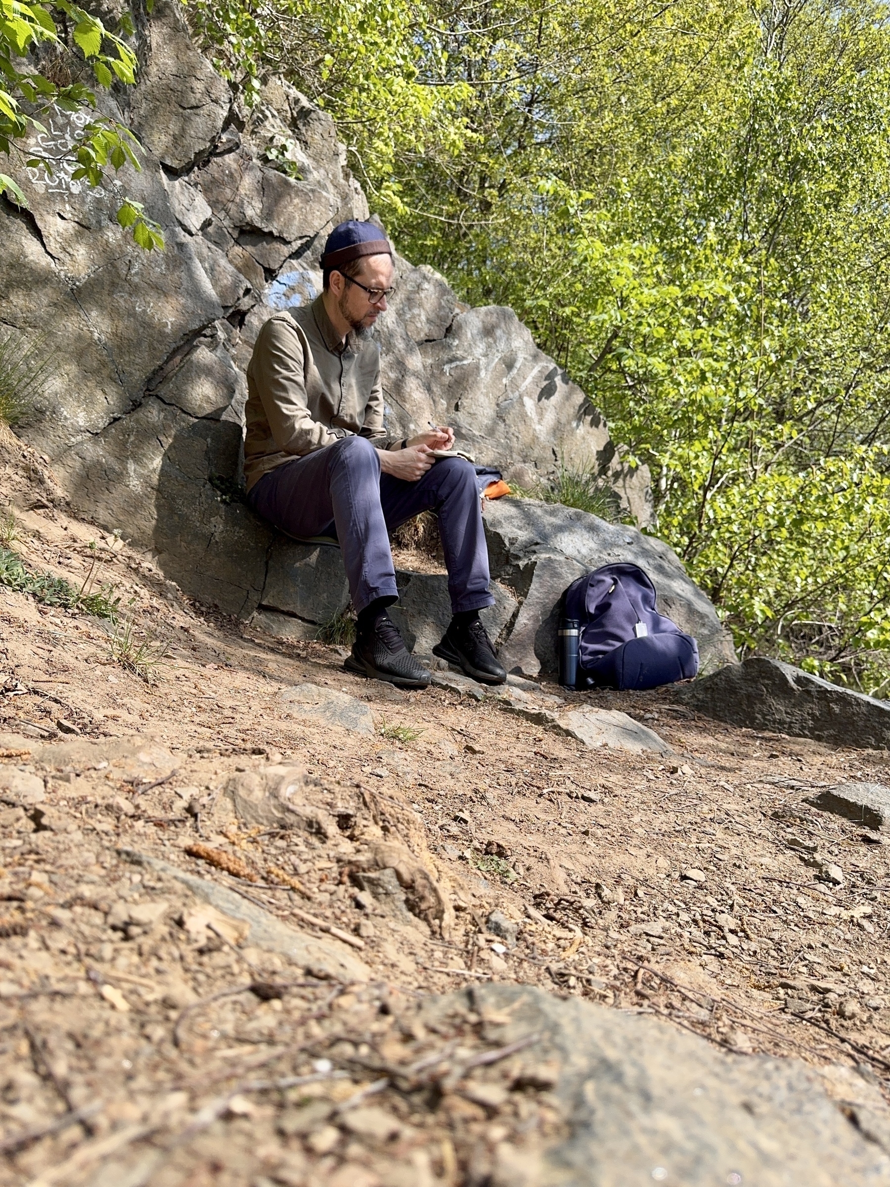 En person sitter på en sten i en skog och tittar på sin mobiltelefon med en ryggsäck bredvid sig.