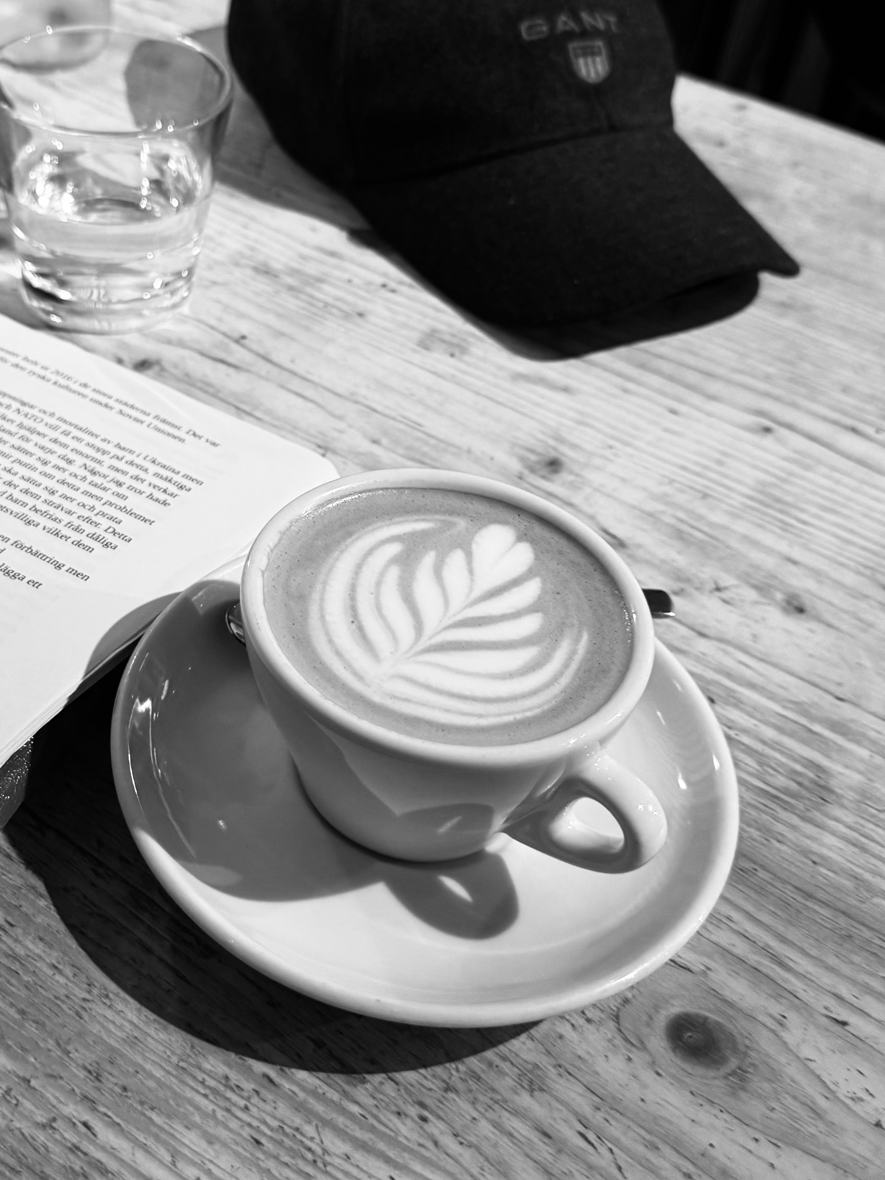 En kopp kaffe med latte art på ett trämaterial bord tillsammans med ett vattenglas, en keps och en öppen bok.
